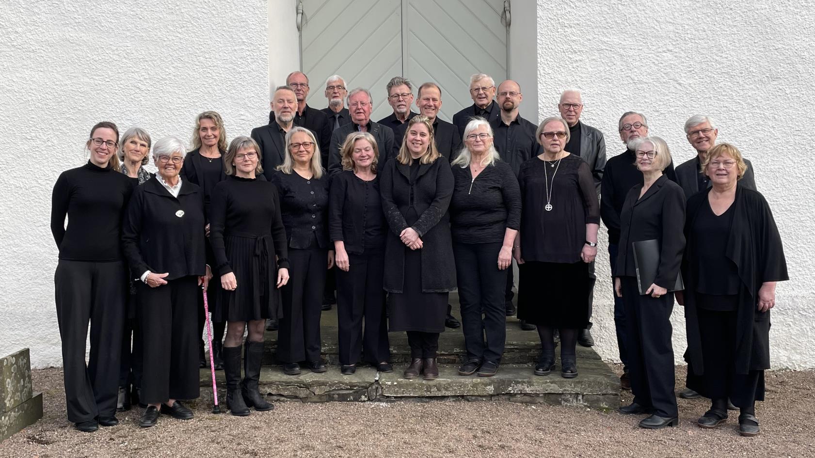 Valla kyrkokör står uppställda framför en kyrkport, alla är klädda i svart