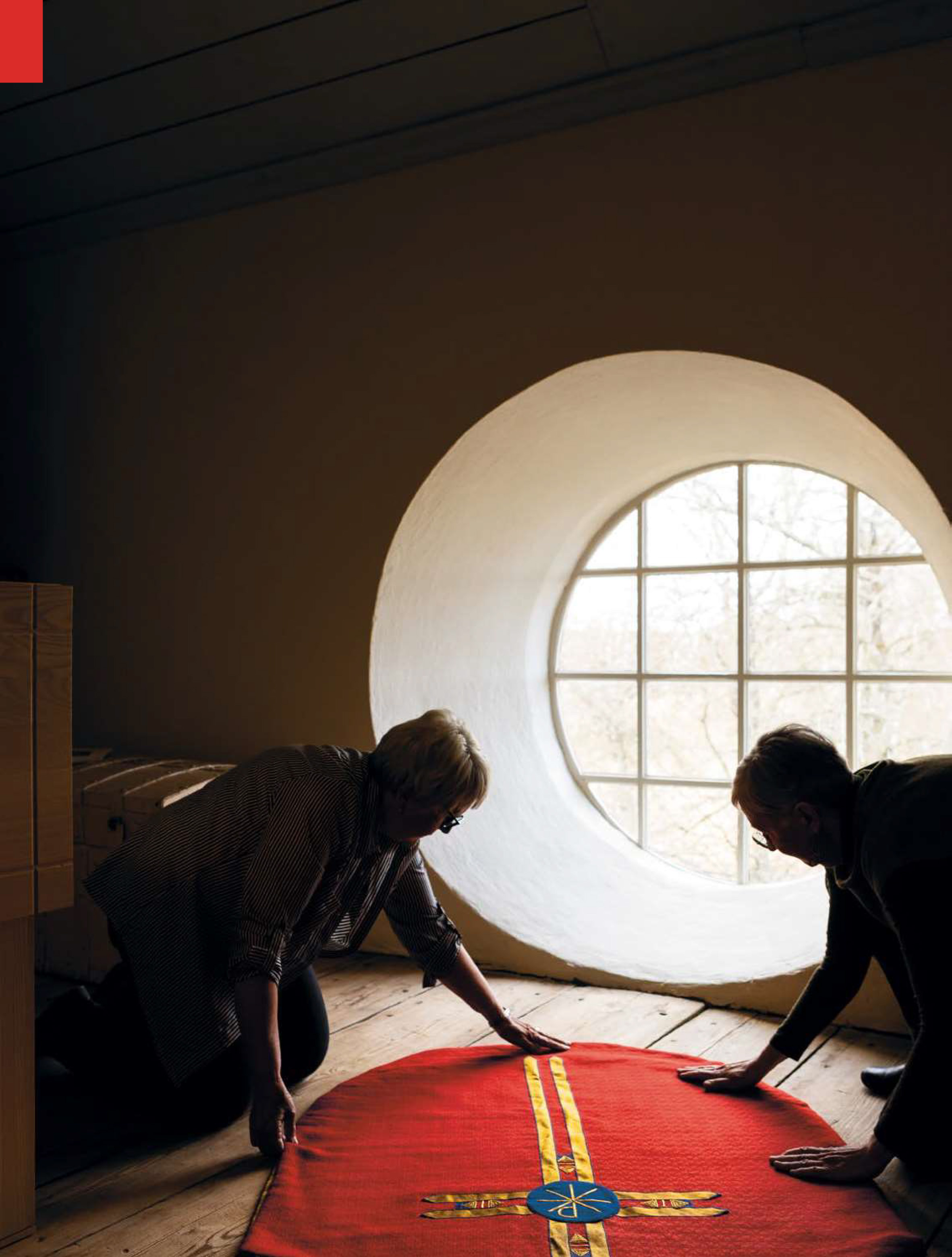 Kvinnor tittar på röd textilier med korsemblem med skarpt dagsljus från runt fönster i Hallingebergs kyrka