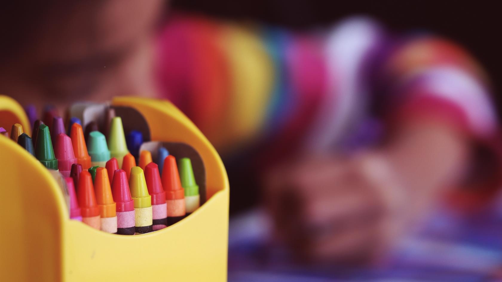 Färgglada målarkritor i en ask. I bakgrunden skymtar ett barn som ritar.