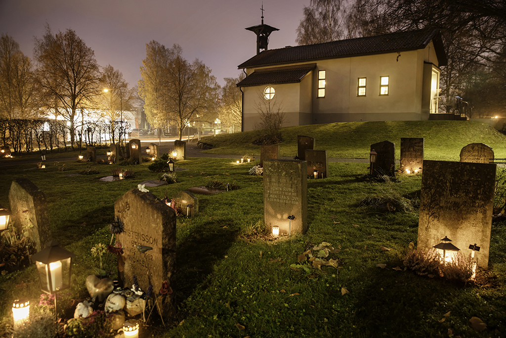 Gravstenar med ljus. I bakgrundne en vit byggnad med kors på taket.