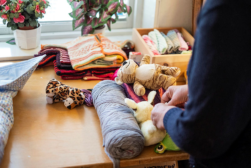 Stickade dockkläder, nallar och garn ligger framme på en bänk vid ett fönster.