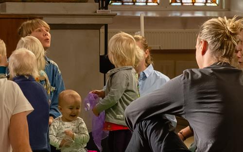 En barngrupp med föräldrar sitter på golvet i Carl Johans kyrka. En lila duk flyger i luften.