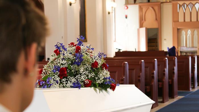 En vit kista med ett stort blomsterarrangemang står i kyrkan. En person i förgrunden kollar på kistan.