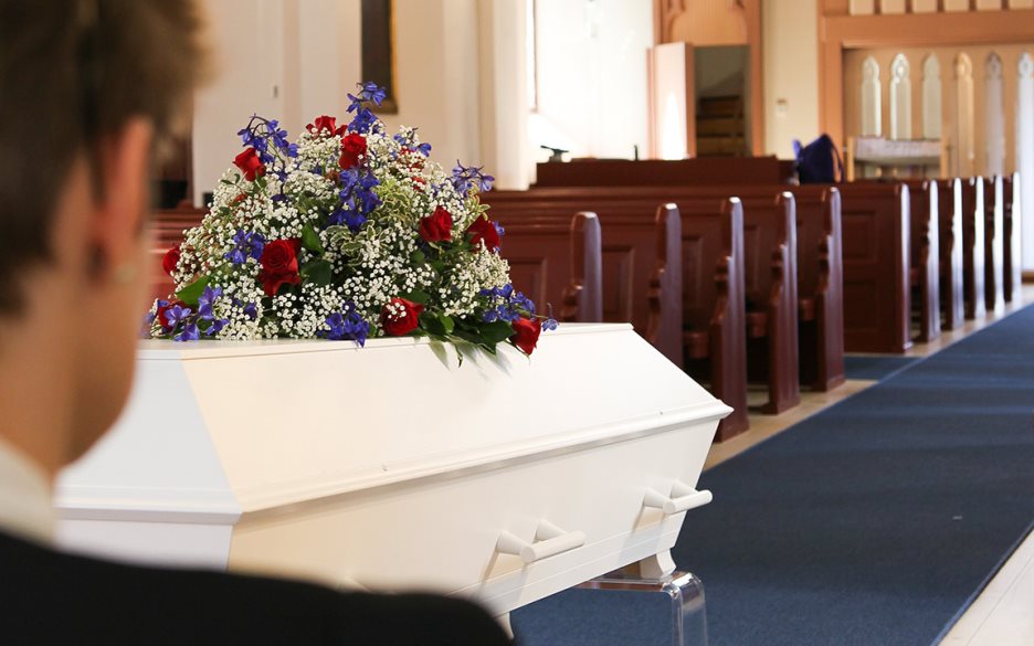 En vit kista med ett stort blomsterarrangemang står i kyrkan. En person i förgrunden kollar på kistan.