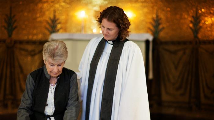 I kyrkan tar en äldre kvinna farväl framför en kista. Prästen står bredvid. Rosa blommor på kistan.