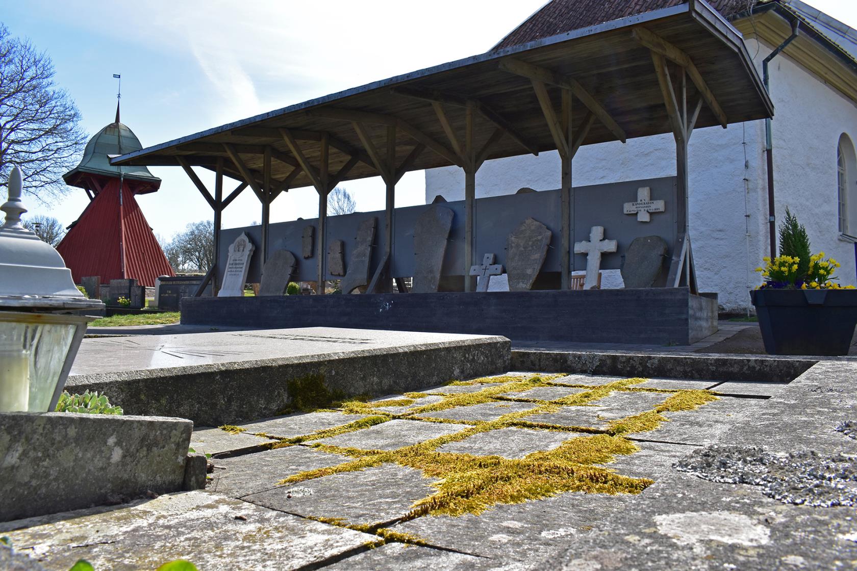 Bergums kyrkogård, lapidariets gamla gravstenar uppställda under tak  med kyrkan i bakgrunden