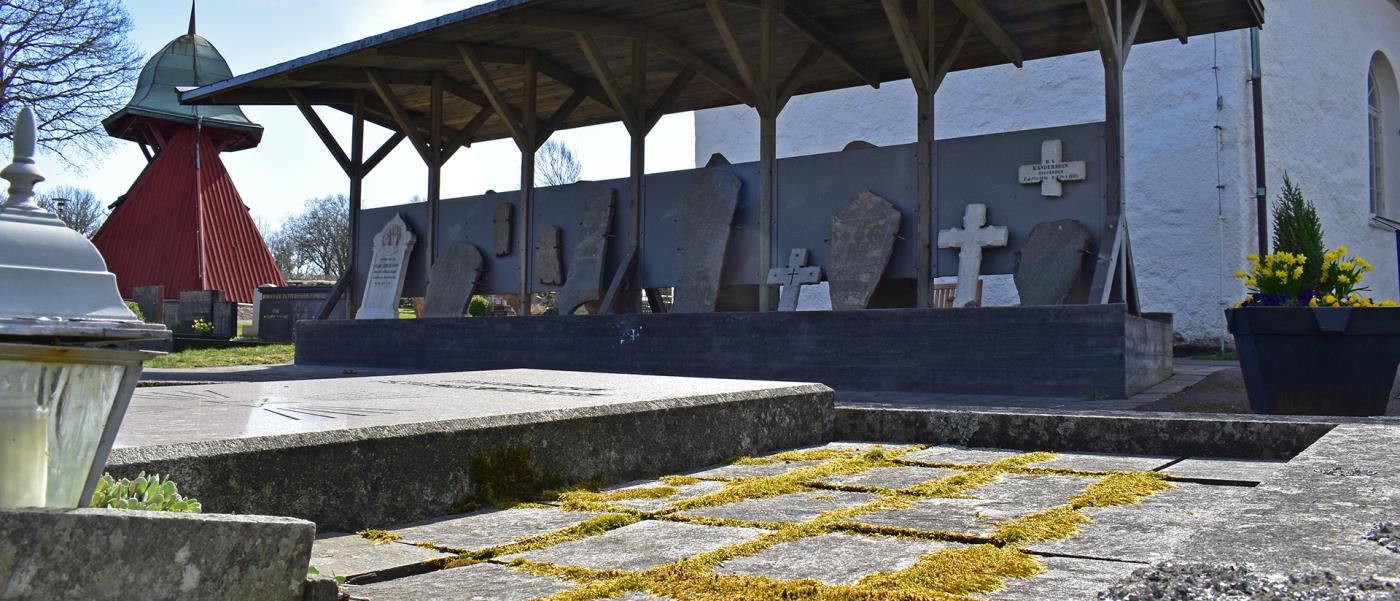 Bergums kyrkogård, lapidariets gamla gravstenar uppställda under tak  med kyrkan i bakgrunden