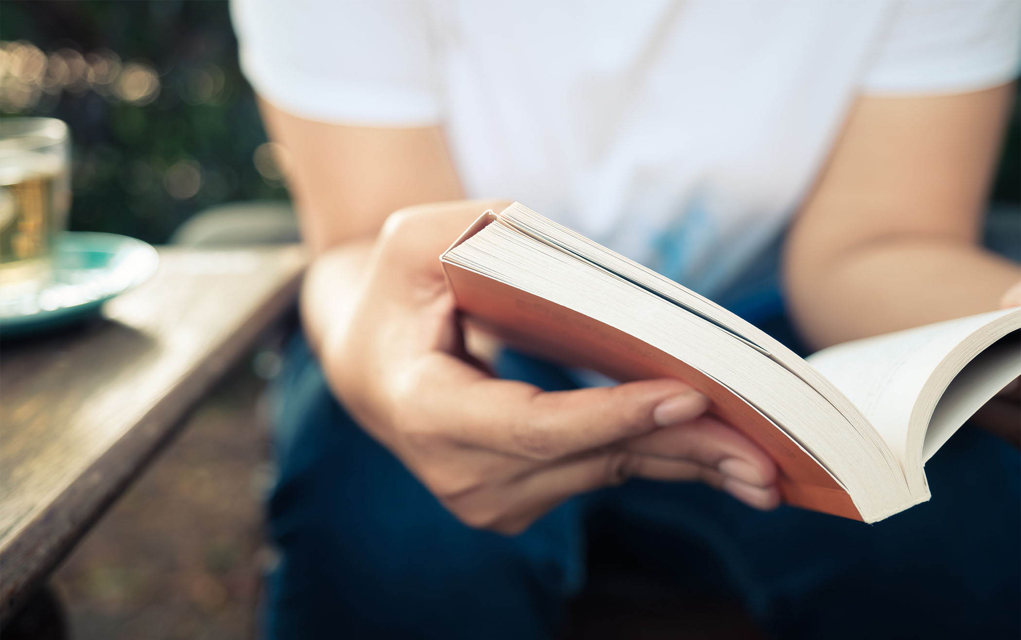 En person i vit tröja håller en bok i sina händer. En kopp står på ett bord vid sidan om. 