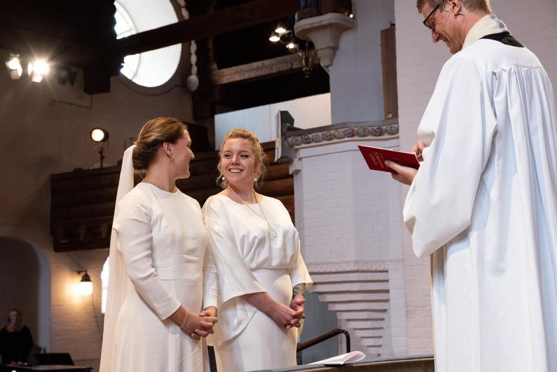 Två kvinnor i brudklänningar ser glada på varandra, står framför prästen. 