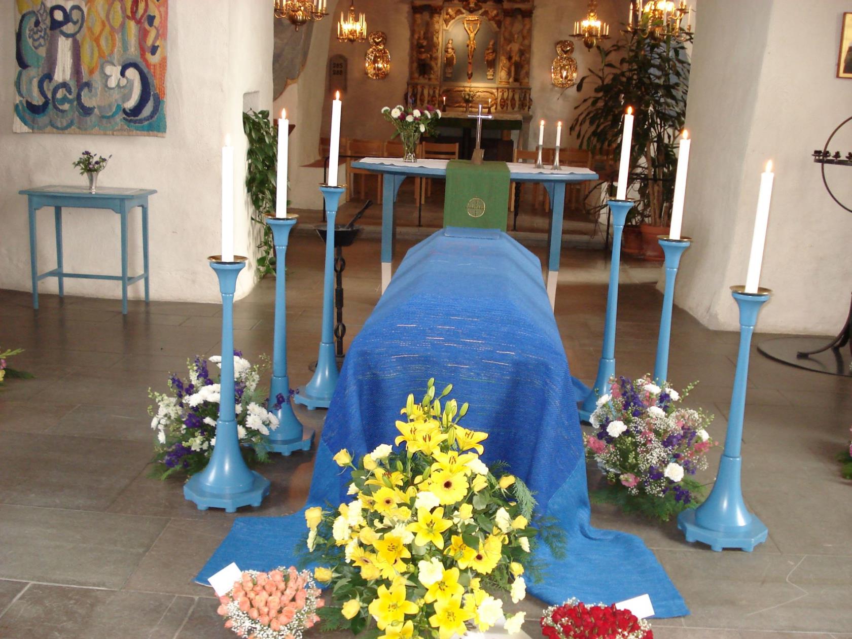Kista i Ödeshögs kyrka. Ovanpå kistan ligger ett blått bårtäcke och och runt kistan står 6 höga träljusstakar med ljus och på golvet runt kistan ligger blombuketter.