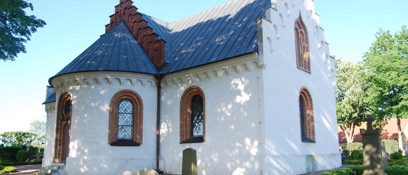 Vitputsad landsortskyrka med rundat kor och röda tegeldetaljer runt fönster och dörrar.