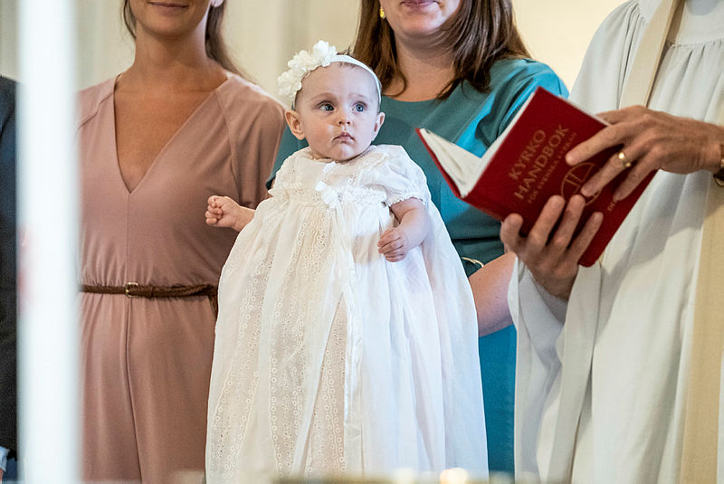 Kvinna i rosa klänning och kvinna i turkos klänning håller barn i dopklänning bredvid präst med bibel.