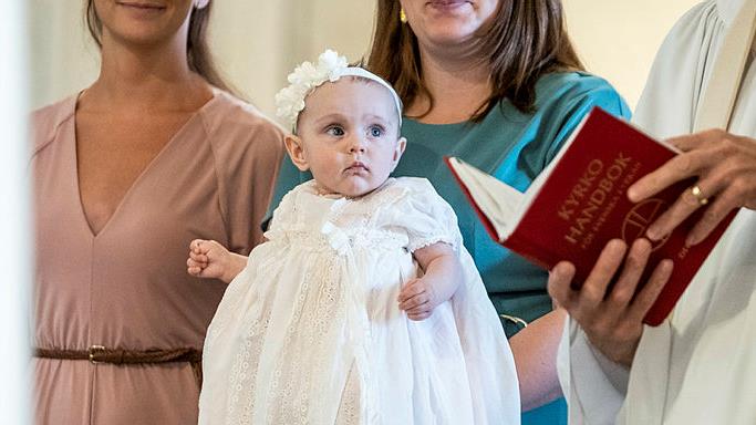 Kvinna i rosa klänning och kvinna i turkos klänning håller barn i dopklänning bredvid präst med bibel.