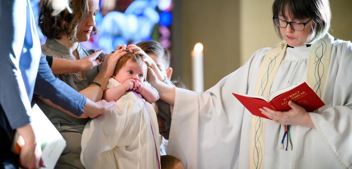 En präst tillsammans med faddrar välsignar ett dopbarn. Liten pojke tittar upp på prästen.