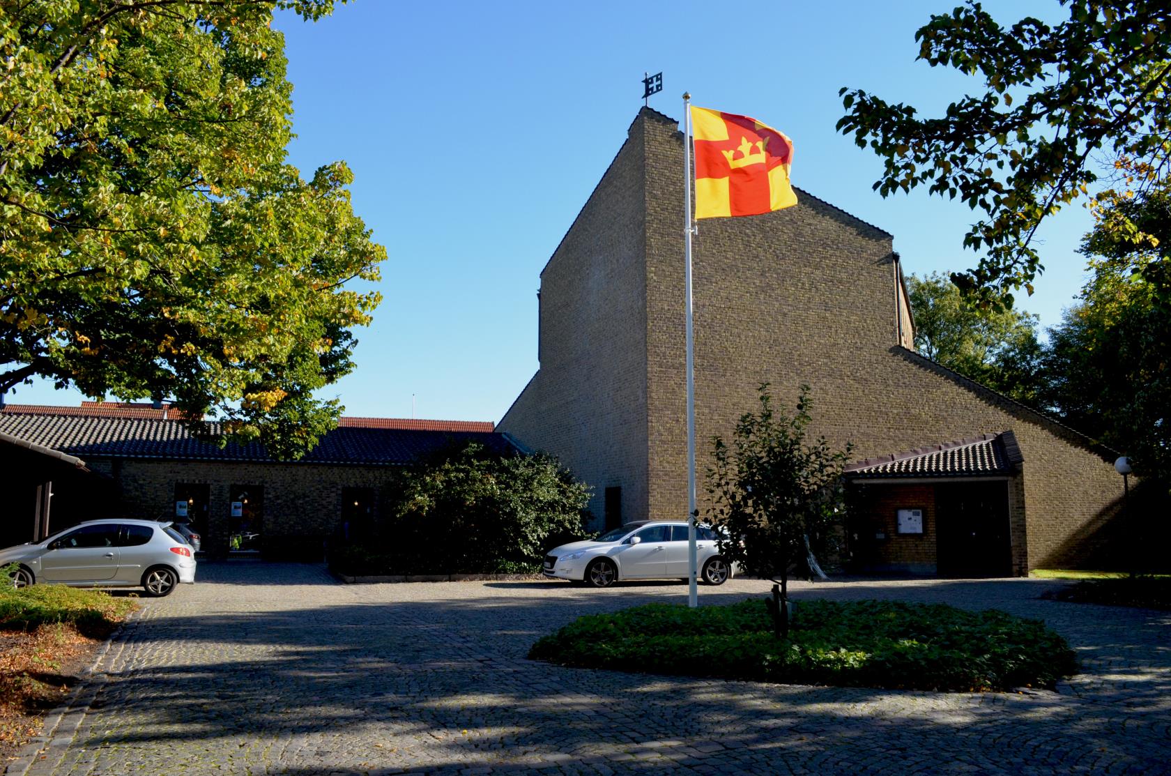 Elinebergskyrkan, en stor byggnad i gult tegel. Själva kyrkobyggnaden är hög och pekar i en spets mot mitten av bilden. Svenska kyrkans flagga i mitten av torget framför kyrkan. Klarblå himmel.