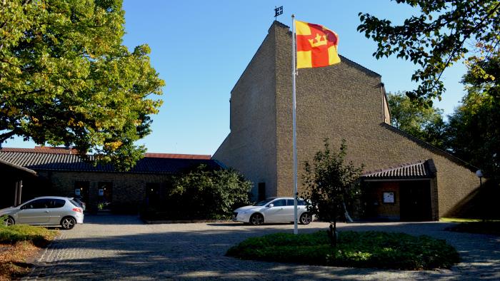 Elinebergskyrkan, en stor byggnad i gult tegel. Själva kyrkobyggnaden är hög och pekar i en spets mot mitten av bilden. Svenska kyrkans flagga i mitten av torget framför kyrkan. Klarblå himmel.