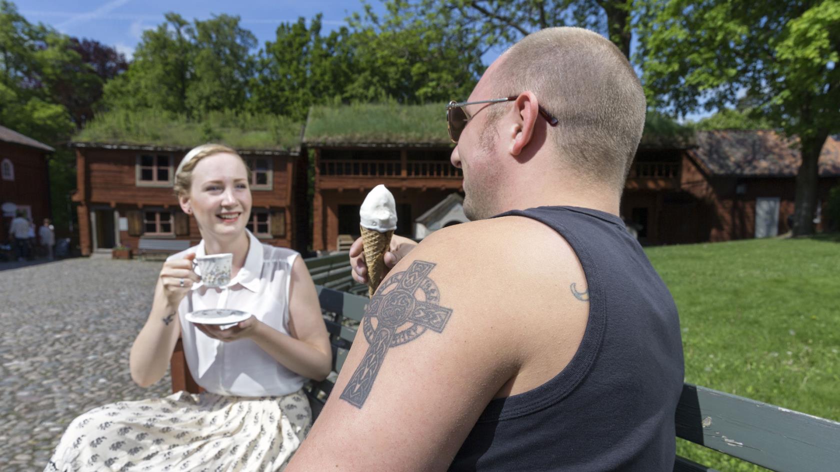 En tjej och en kille fikar i Wadköping. I förgrunden syns killens axel med en tatuering i form av ett kors.