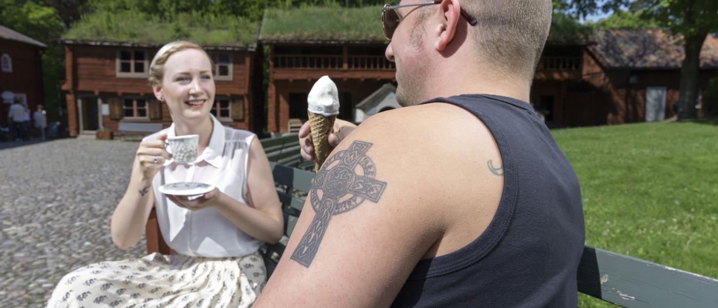 En tjej och en kille fikar i Wadköping. I förgrunden syns killens axel med en tatuering i form av ett kors.