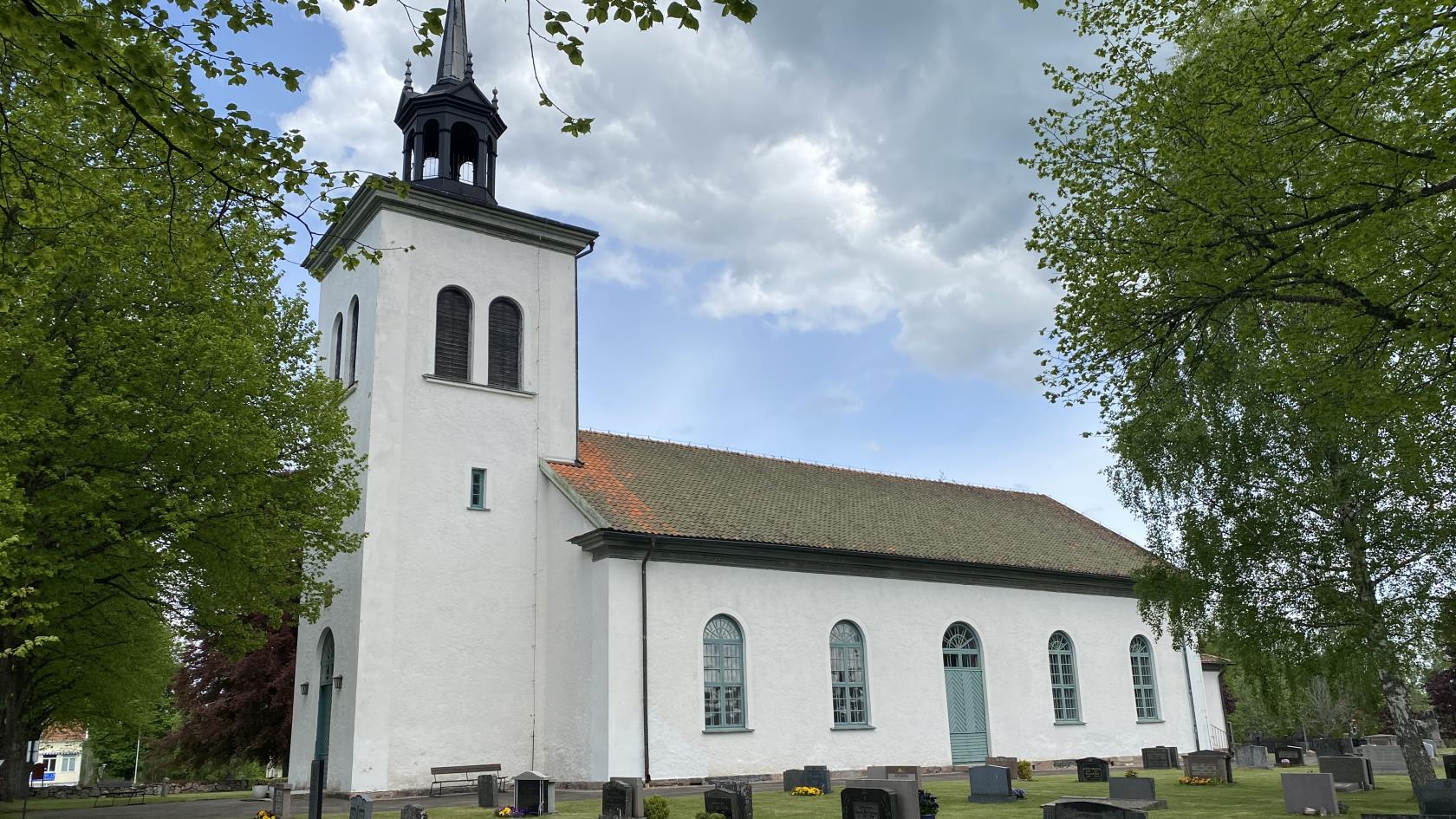 Fröjereds kyrka från sidan, med torn och långhus, framför syns kyrkogården med gravstenar och grönt gräs på marken, träden är lummigt gröna och himlen är blå med några moln.