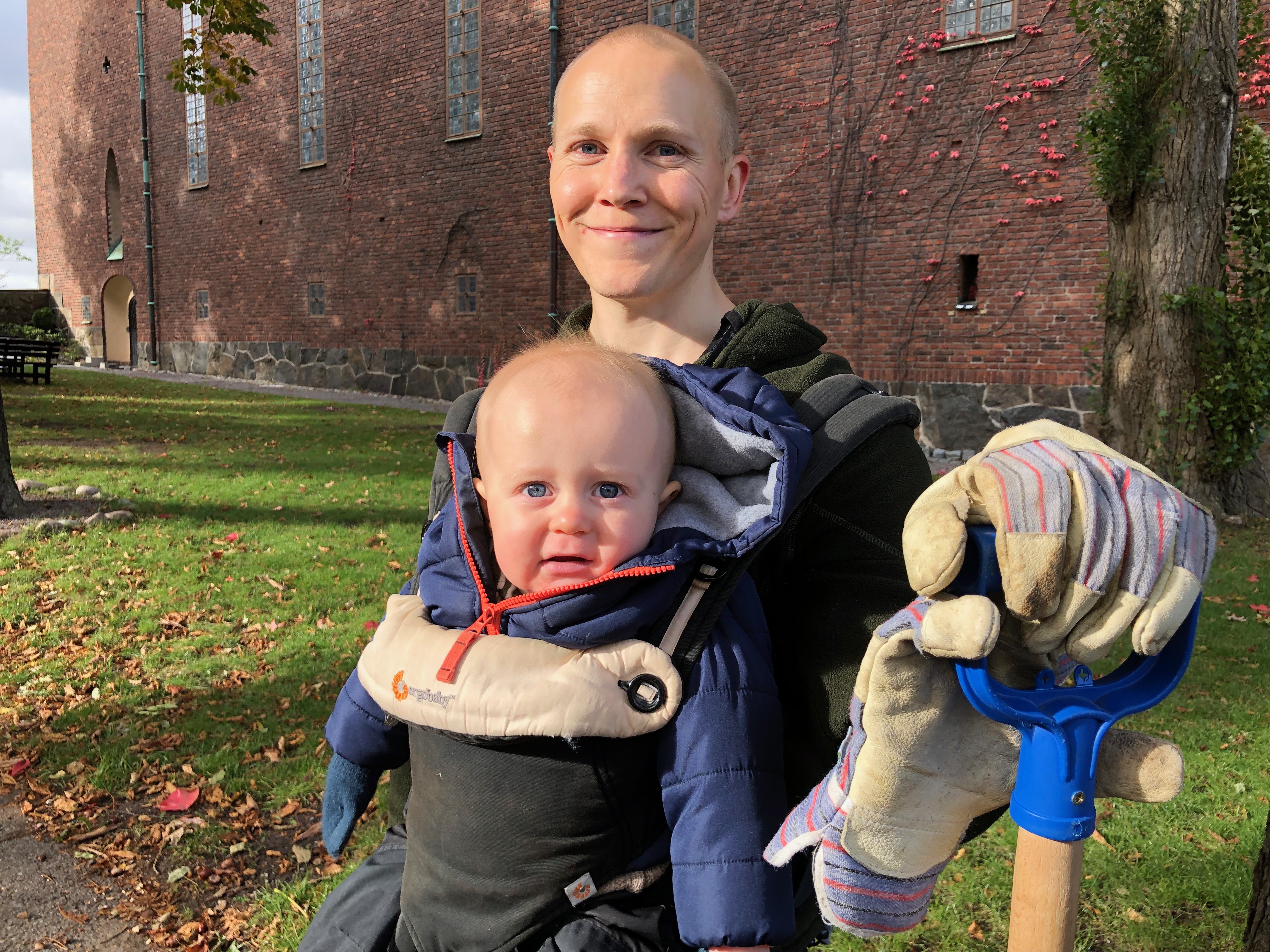 En person sitter och har en bebis i sitt knä och ett spadhandtag i ena handen som är i en handske. Det är en stor byggnad, samt gräs och löv i bakgrunden.  