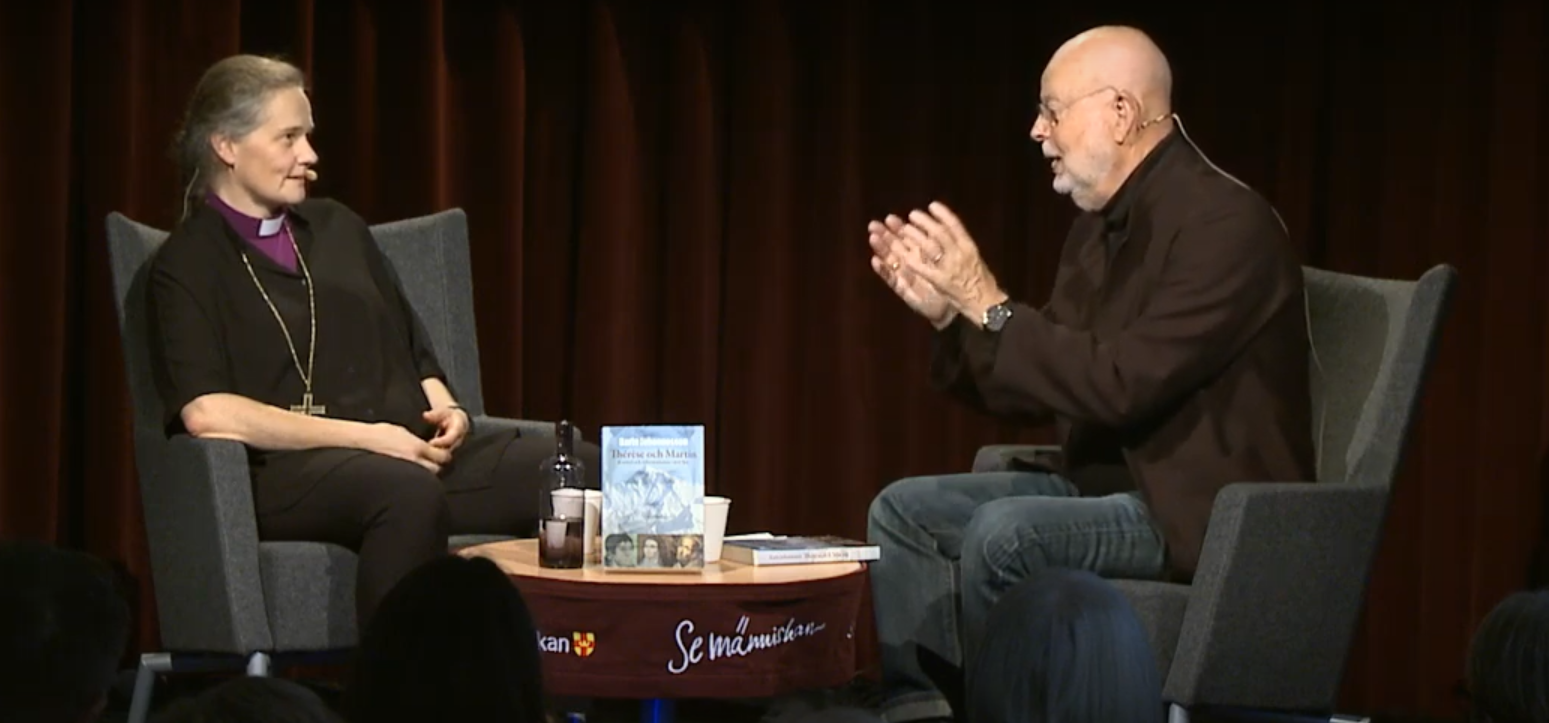 Två personer sitter i varsin fåtölj på en scen.