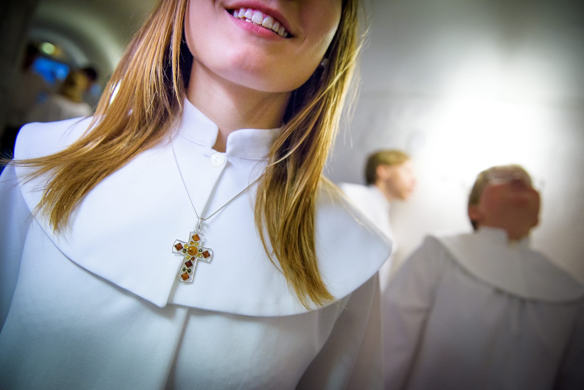 Nedre delen av ansiktet hos en leende konfirmand. Långt ljust hår. Personen är klädd i en konfirmandkåpa och har ett halsband med ett kors på sig. I bakgrunden kan man skymta andra konfirmander. 