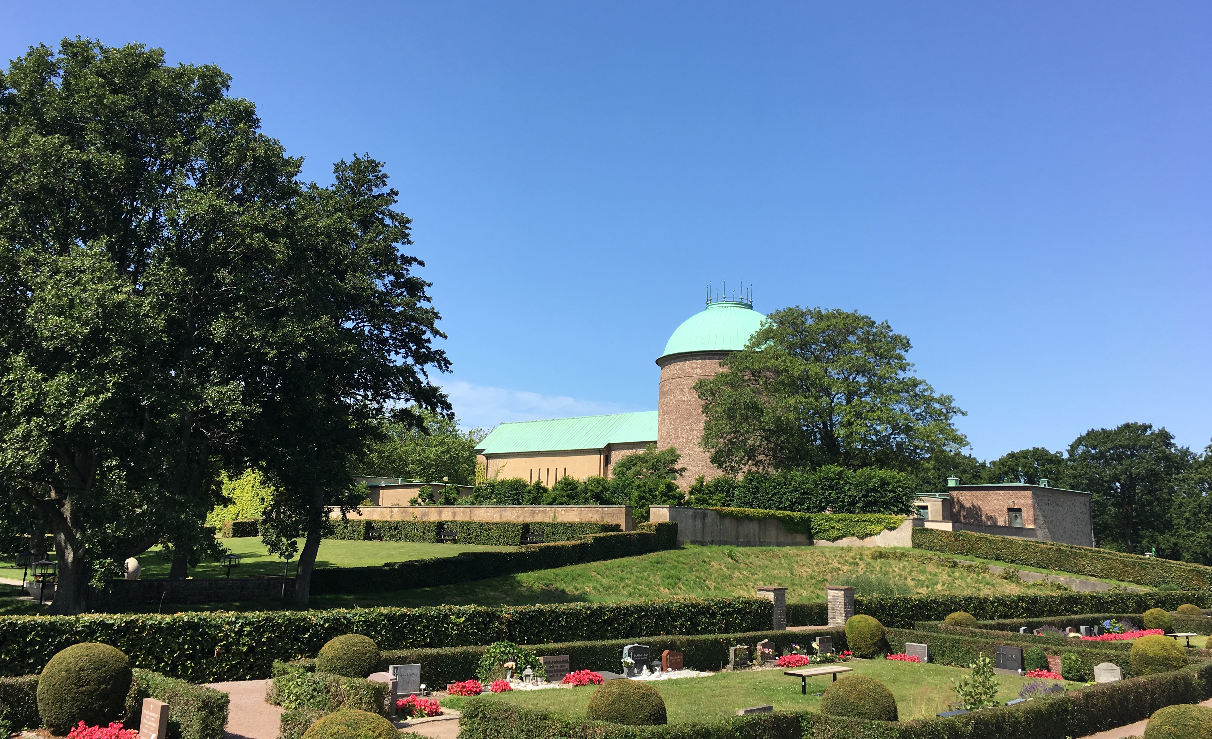 Kyrkogård med gravstenar, träd, och annan grönska. Ett krematorium från 1920-talet med runt torn finns i bakgrunden.