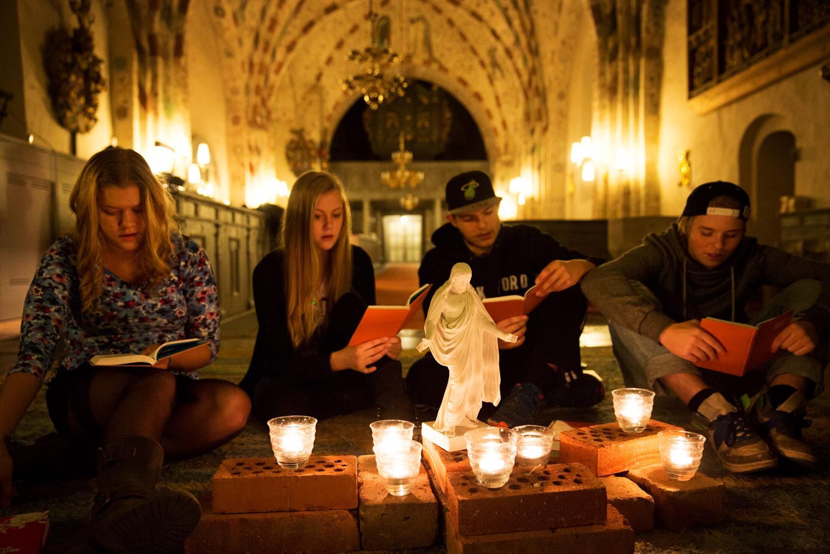 Ungdomar sjunger sittande på golvet i kyrka. Tända ljus och Jesusstaty framför