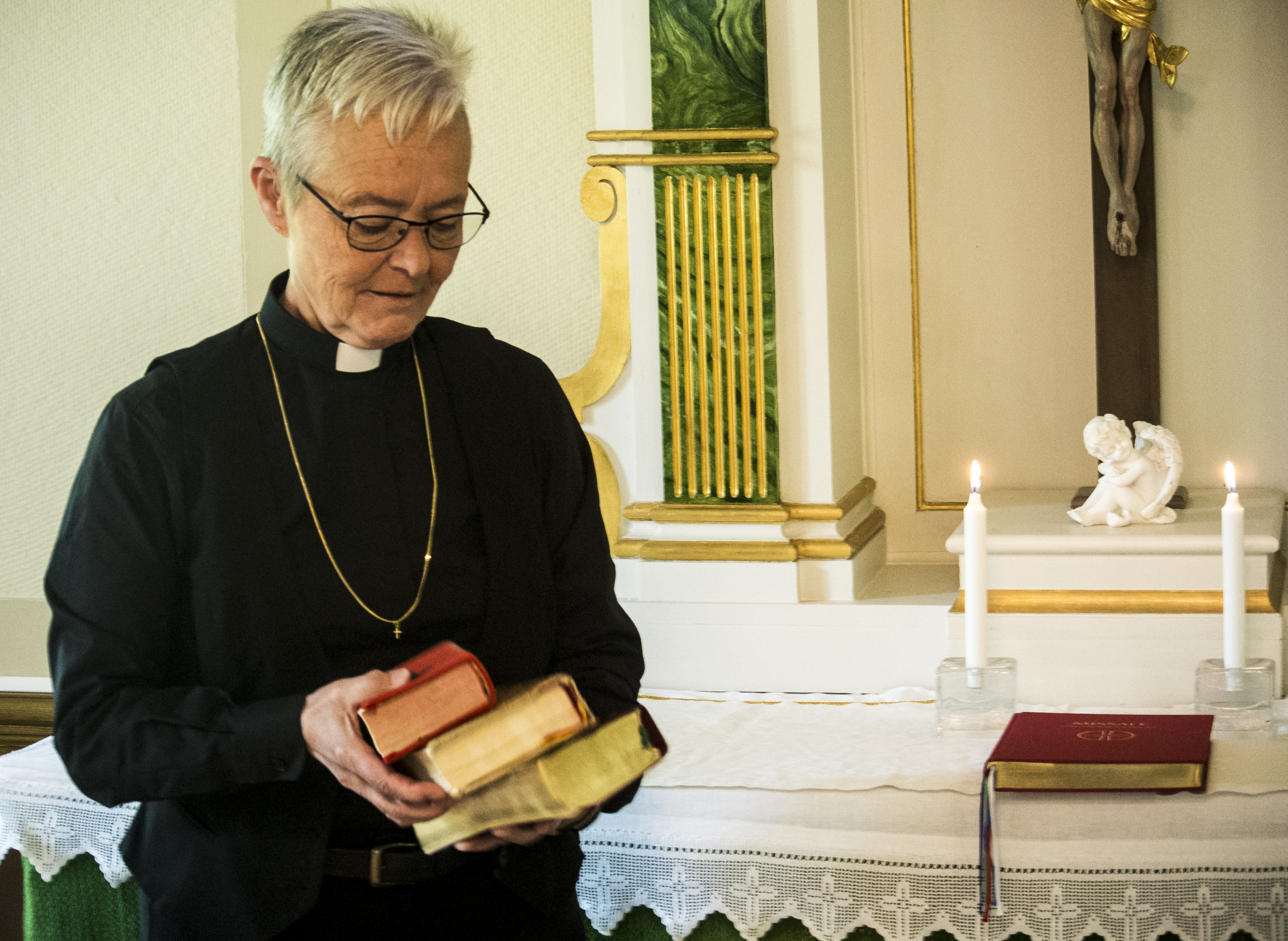 En äldre kvinna som är präst står i Karlholms kyrka. Hon har kort, blont hår, svart prästklädsel, vit krage, guldkedja runt halsen och håller i en trave böcker. Hon står vid kyrkans altare.