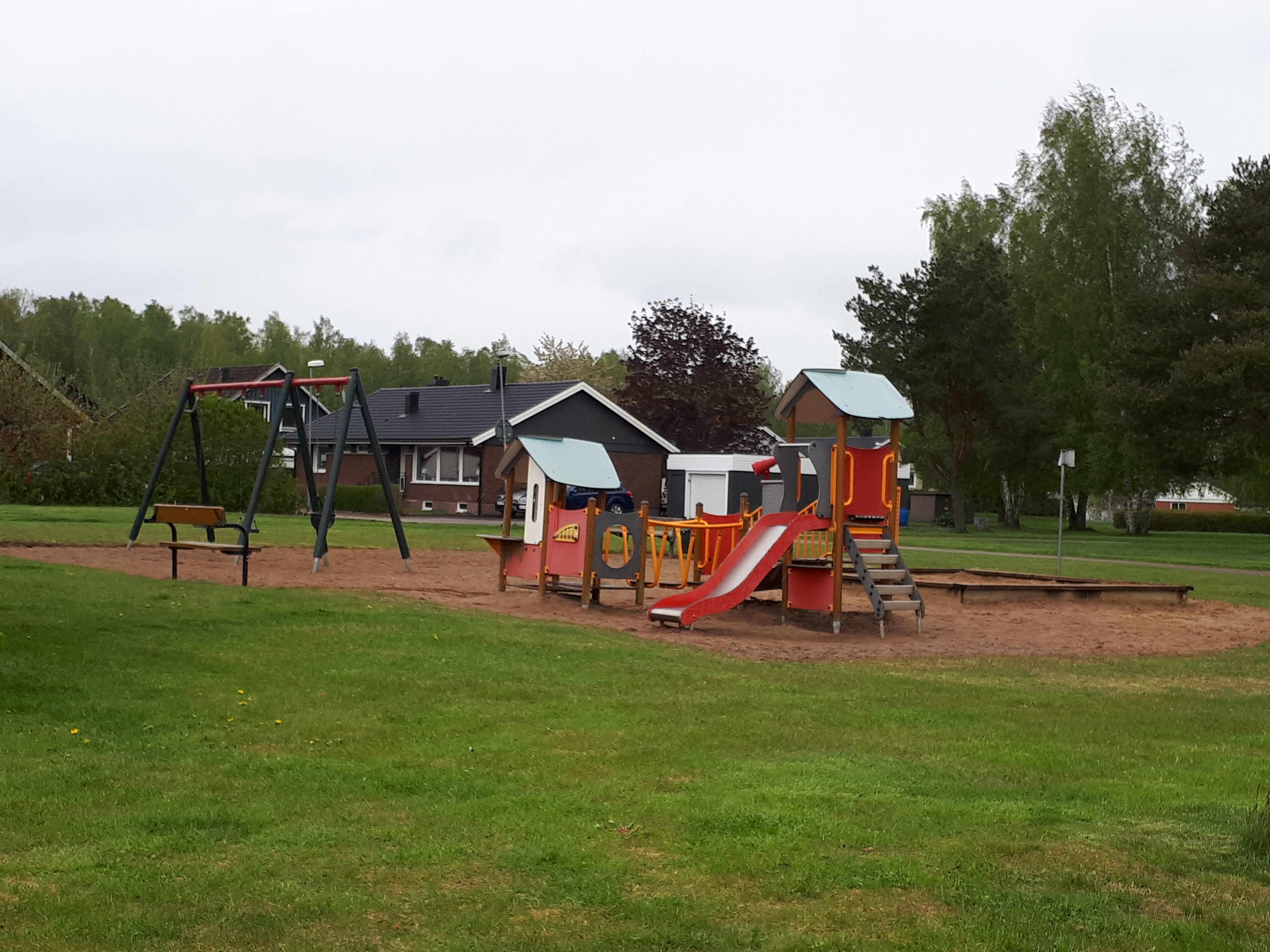 En lekpark med gungställning, rutschkana, små lekhus med gångbro i mellan husen och sandlåda. I bakgrunden syns villor.
