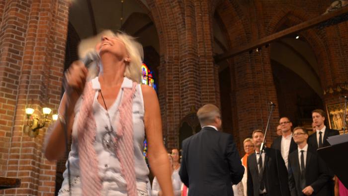 Sångerska i förgrunden sjunger gospel. Kör syns i bakgrunden. I bakgrunden syns också höga valv i rött tegel. Medeltida kyrka.