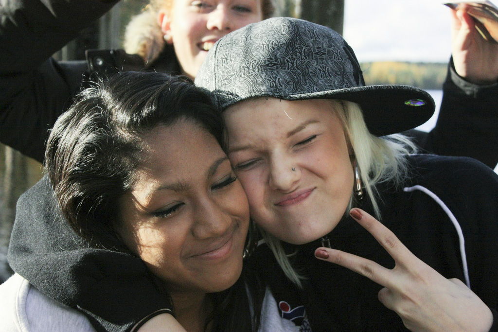 Två tonårstjejer kind mot kind med ansikten mot kameran. De blundar, grimaserar på skoj, gör V-tecken.