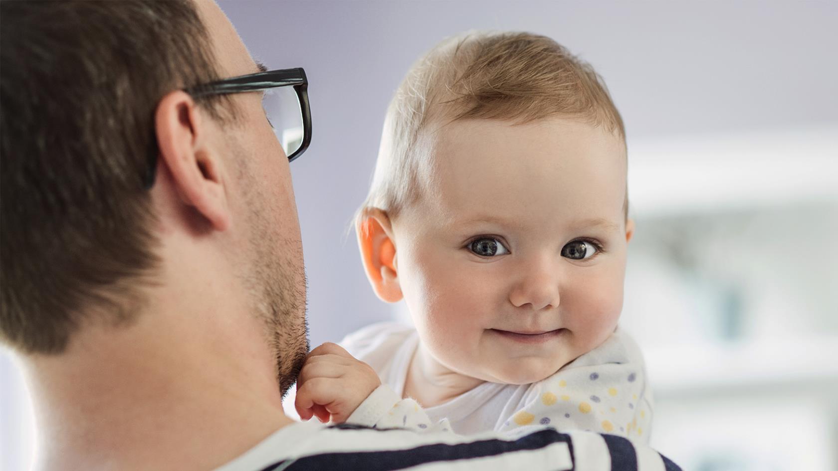 En person i glasögon håller i en bebis som ser glad ut. 