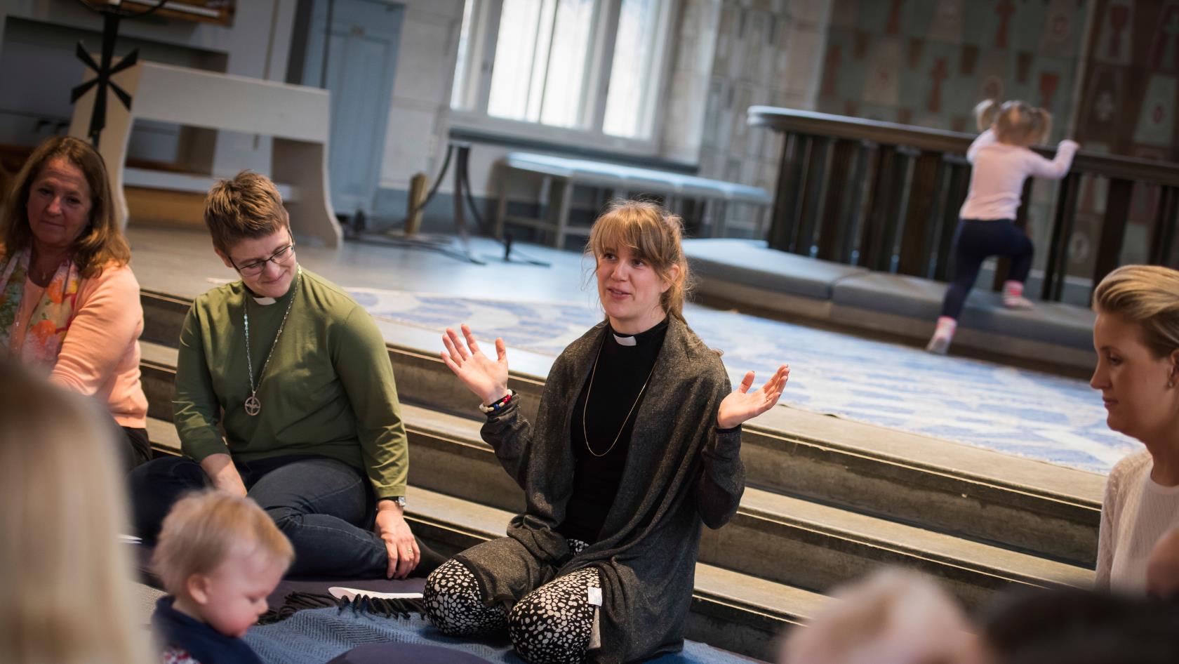 En präst leder en samling med små barn och deras föräldrar. De sitter på golvet i en ring nedanför altaret i en kyrka. En liten flicka försöker klättra över altarringen i bakgrunden.  