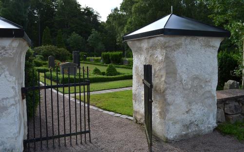 En svart järngrind står lite öppen. I bakgrunden skymtar kyrkogården.