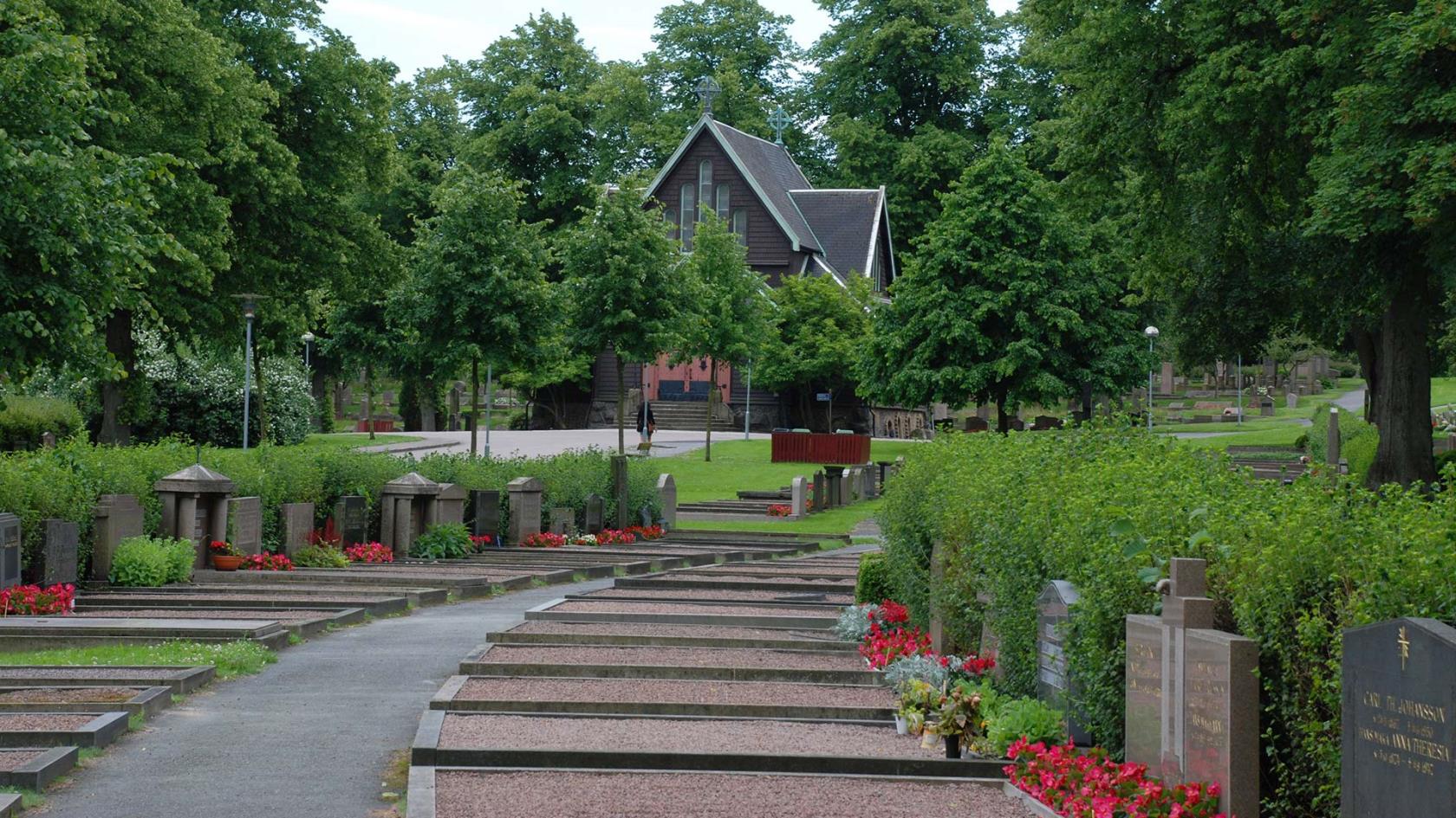 En lummig kyrkogård med gravar i förgrunden och ett kapell i bakgrunden.