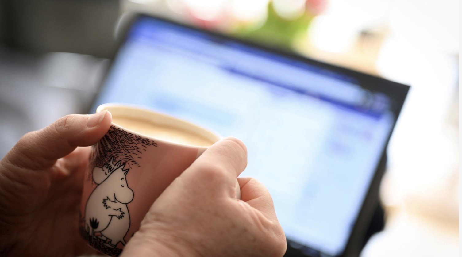 En mugg hålls av två händer och i bakgrunden syns en datorskärm. 