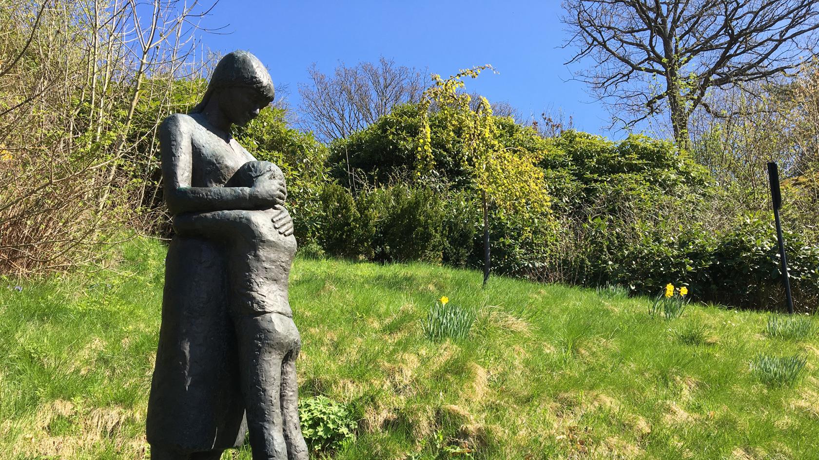 En vuxen person omfamnar ett barn. Skulptur i grässlänten ovanför minneslunden med buskar och träd i bakgrunden