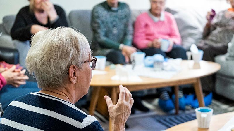 Äldre kvinna i randig tröja samtalar med andra män och kvinnor.