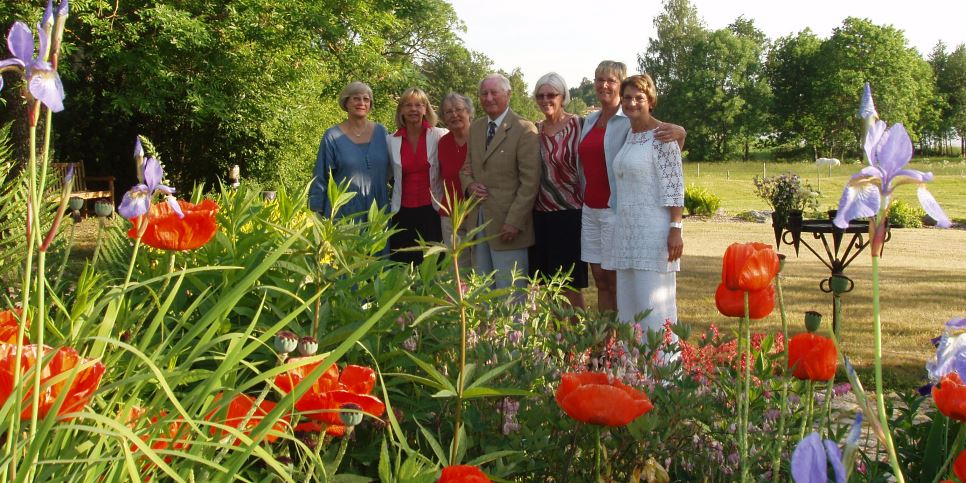 Några personer står samlade på en gräsmatta en vacker sommardag, framför finns blommor och bakom finns träd