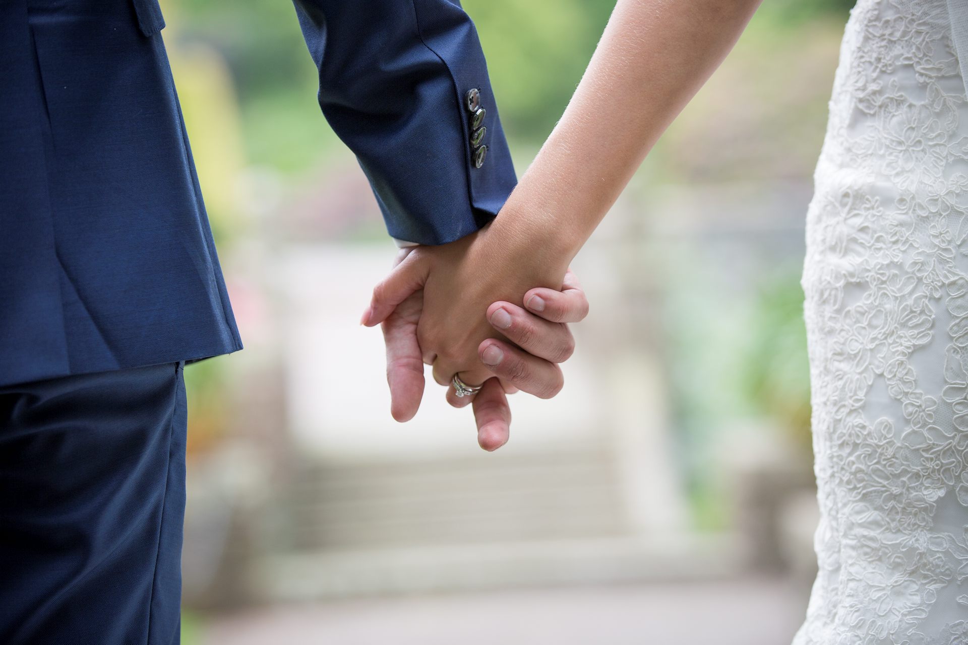 Brudgum i blå kostym och brud i vit klänning håller varandra i händerna.