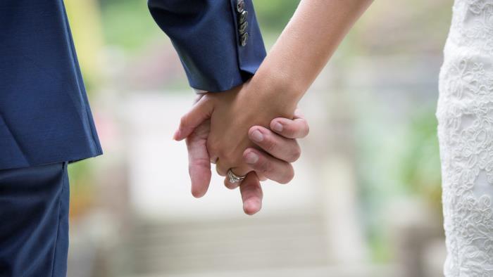 Brudgum i blå kostym och brud i vit klänning håller varandra i händerna.