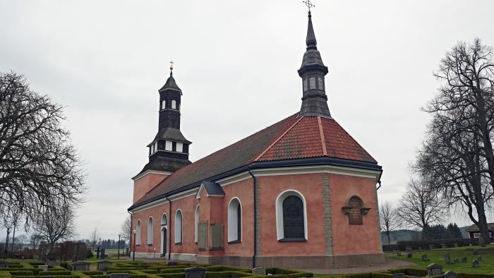 Ekeby kyrka med gravar omgärdade av häckar i förgrunden. Kyrkan är röd med tegeltak.