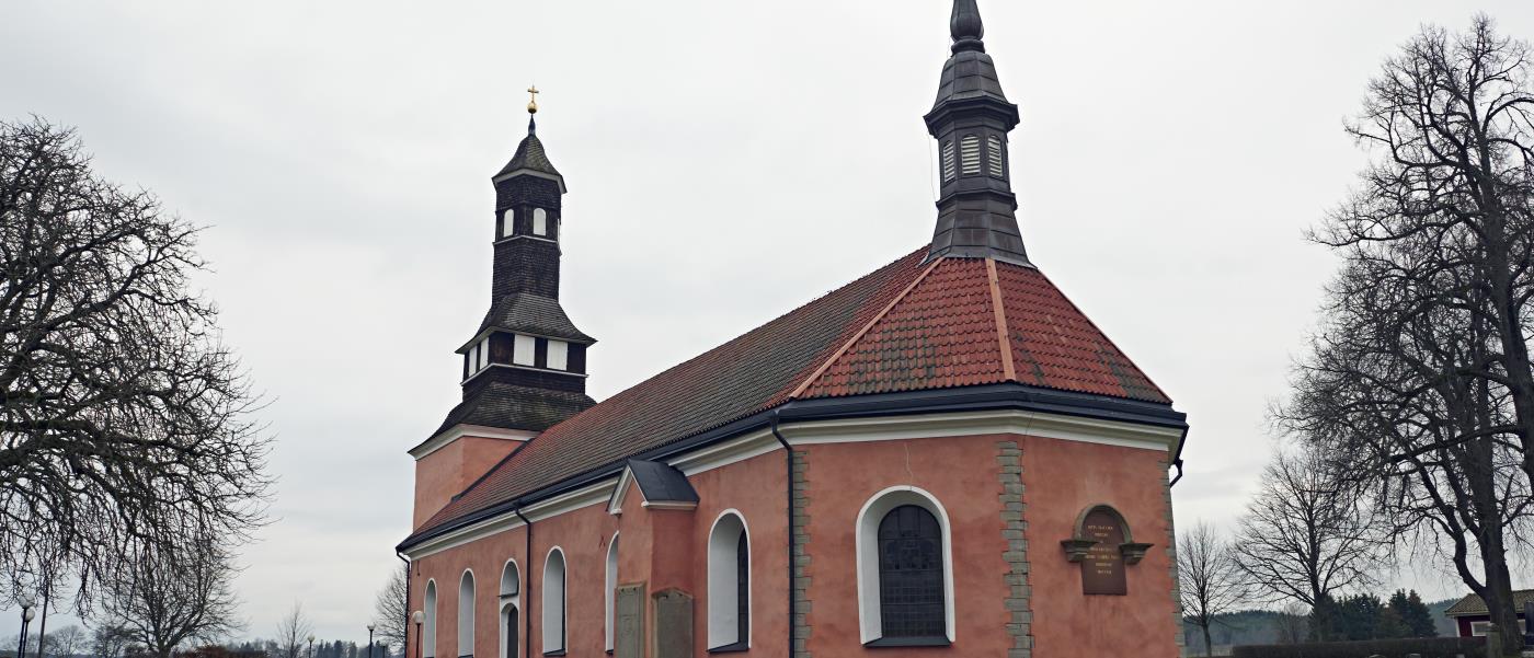 Ekeby kyrka med gravar omgärdade av häckar i förgrunden. Kyrkan är röd med tegeltak.