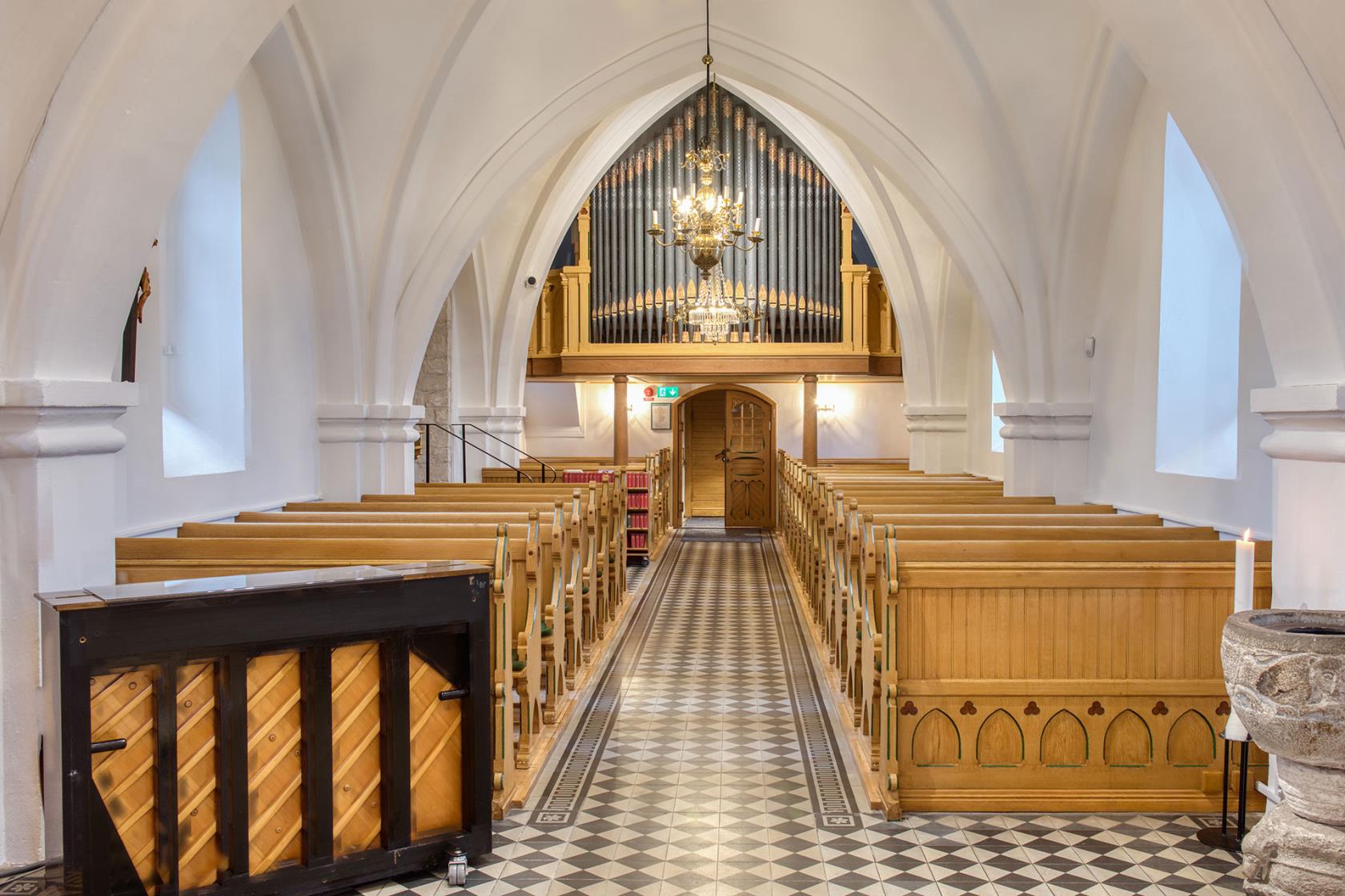 Vitkalkat kyrkorum med ljus bänkinredning i trä, orgelläktare och dopfunt i sandsten.