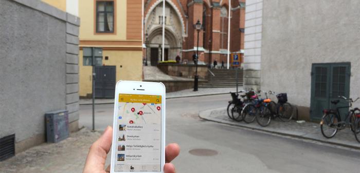 Hand håller i mobiltelefon med appen på skärmen, framför Uppsala domkyrka.
