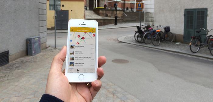 Hand håller i mobiltelefon med appen på skärmen, framför Uppsala domkyrka.