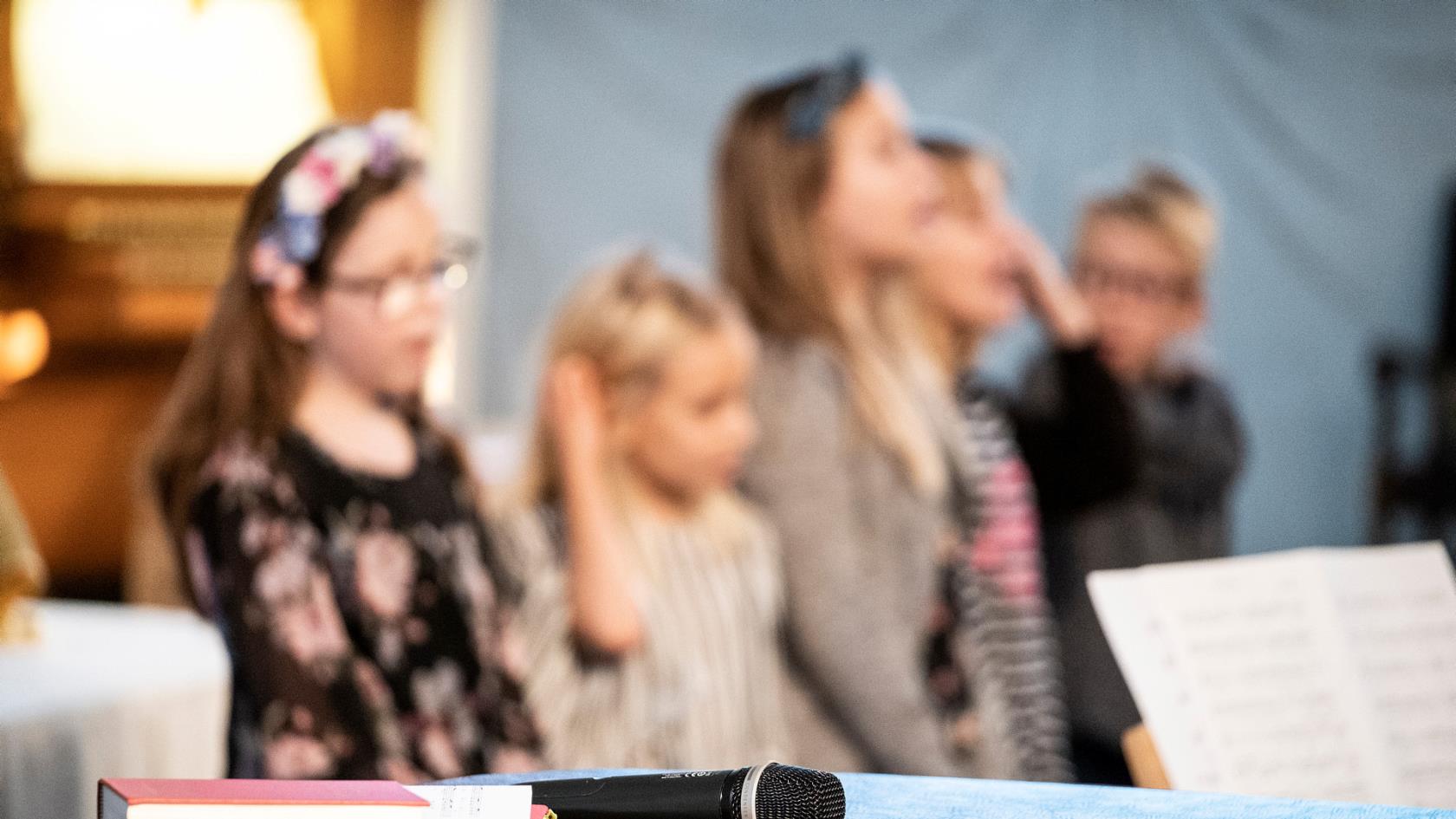 En psalmbok och en mikrofon ligger på ett bord. Bakom sjunger några barn.