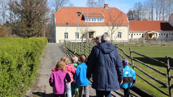 En grupp med barn som går längst vägen tillsammans med två vuxna. Barnen har olika färger på sina jackor och solen skiner. I bakgrunden syns ett stort gult hus som barnen och de vuxna rör sig mot.