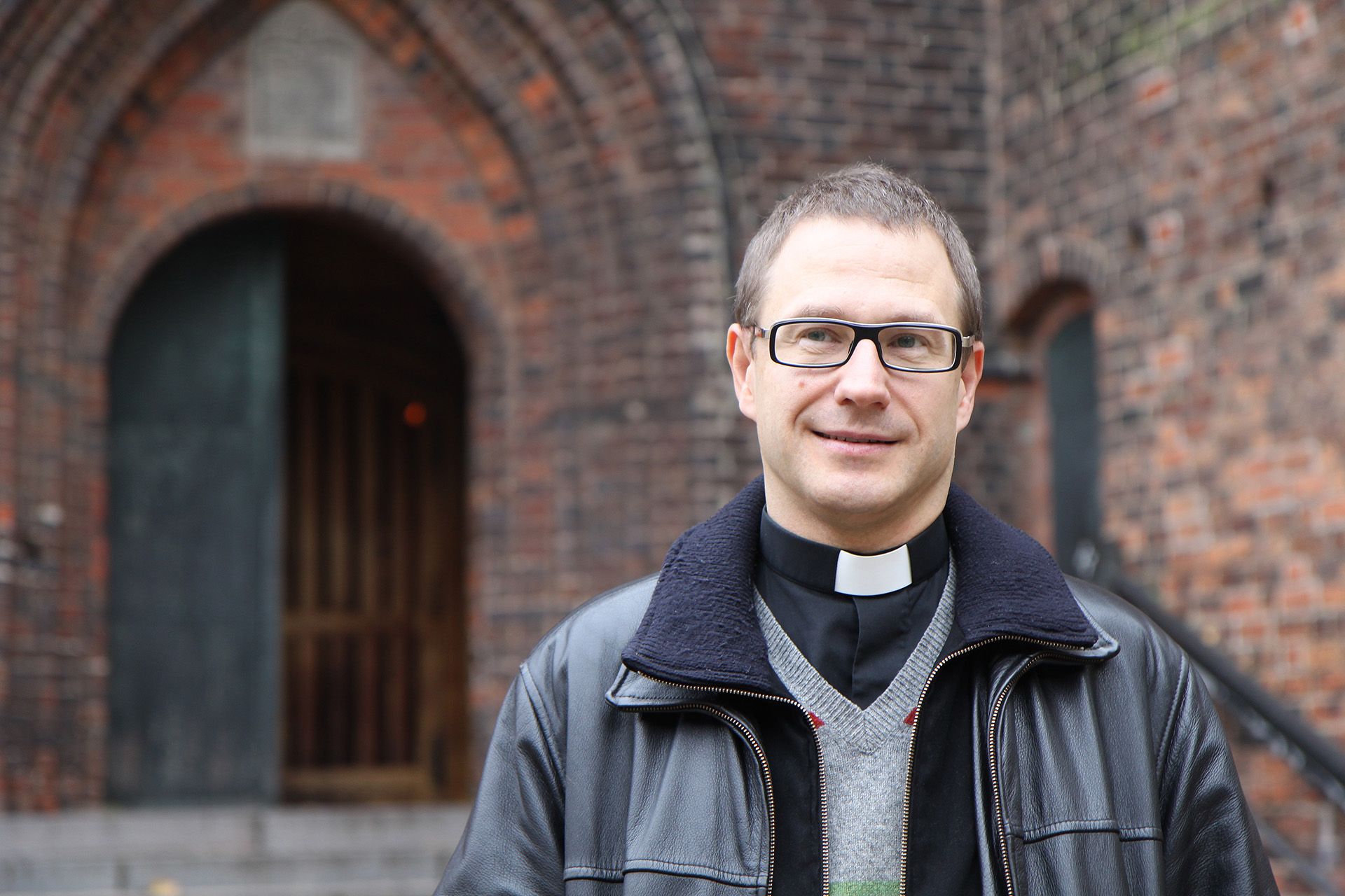 Porträtt av en manlig präst iklädd prästskjorta och skinnjacka. Röd medeltida tegelkyrka i bakgrunden.